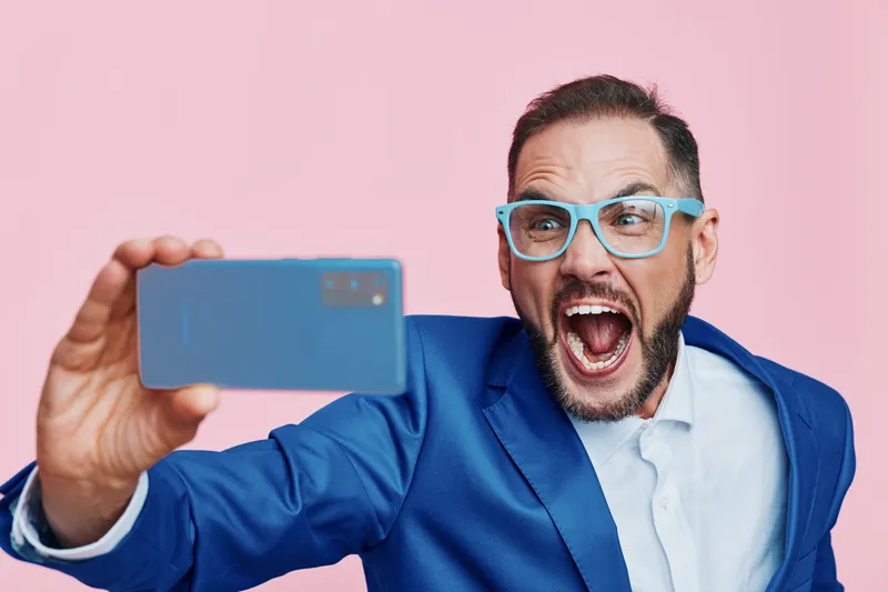 A professional man in a blue suit takes a dynamic selfie against a solid pink background, conveying strong emotion, energy, and confidence in a modern business moment and ready for promotion.
