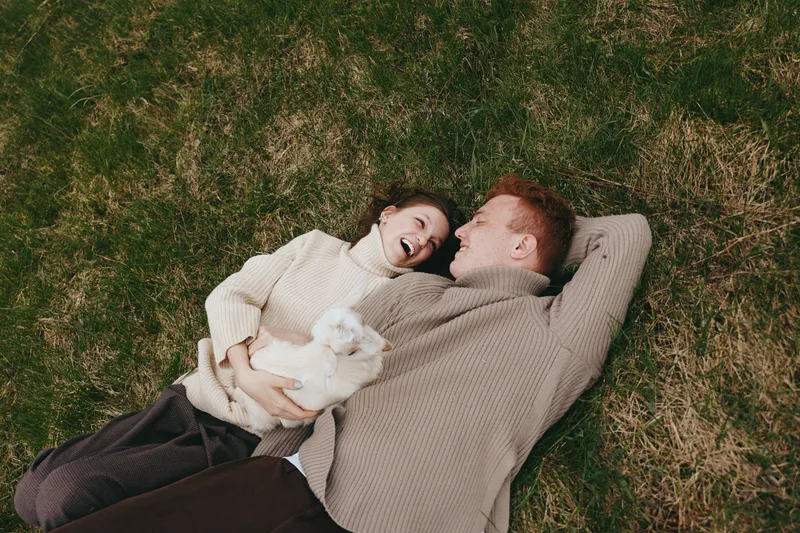 Smiling couple lying on grass in sweaters, relaxing together with a small white dog between them, enjoying a cozy outdoor moment, warm and tender lifestyle scene. Together they gaze softly at each