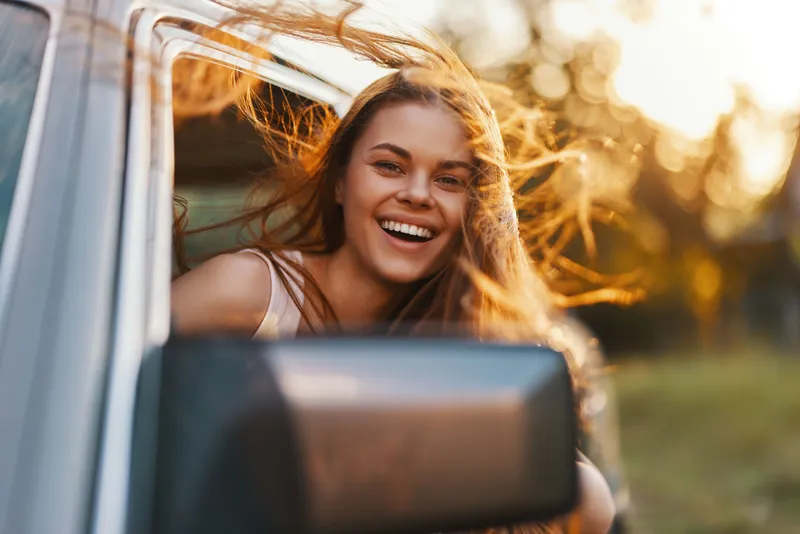 Smiling young woman with flowing hair enjoying a road trip in a car during sunset, capturing the essence of freedom and adventure.