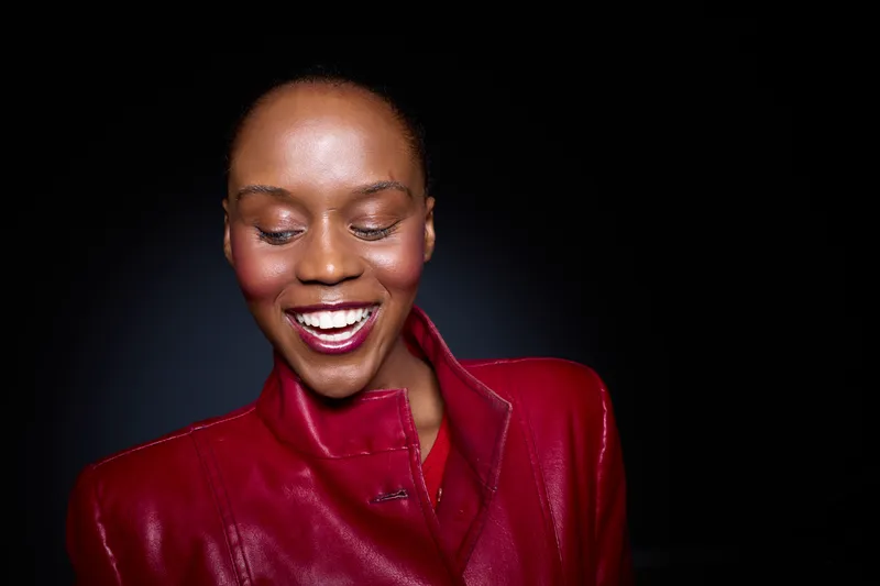Smiling African woman with dark skin wearing a red leather jacket looks down with eyes closed against a black background. Concept of happiness, fashion, and beauty with natural expression.
