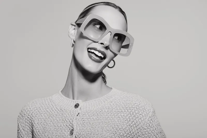 Smiling slender white woman with slicked hair and oversized sunglasses wearing textured sweater on light background.
