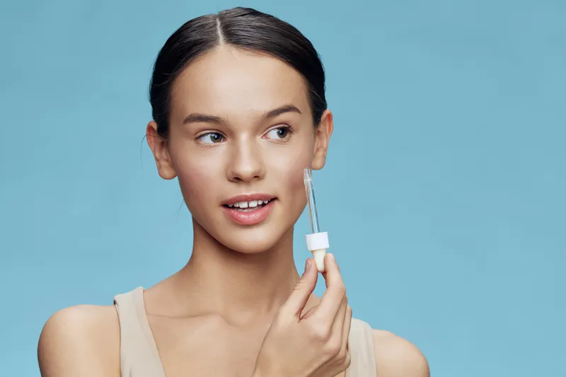 Young woman applying serum on her face, showcasing radiant skin against a light blue background, expressing confidence and beauty in a skincare routine