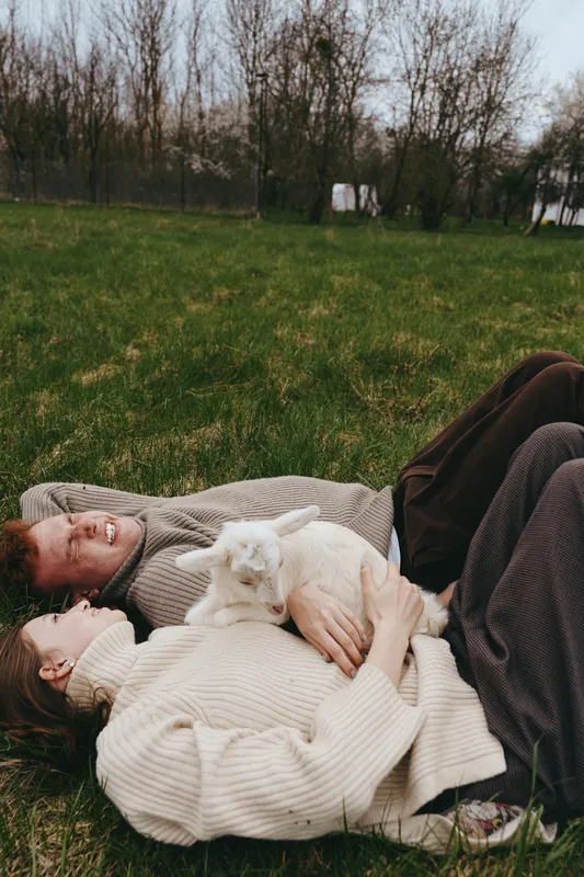 Relaxed couple lying on a blanket in a grassy park with a white goat kid cuddling between them, conveying warm togetherness, nature calm, gentle companionship, inspiring mindfulness and well being