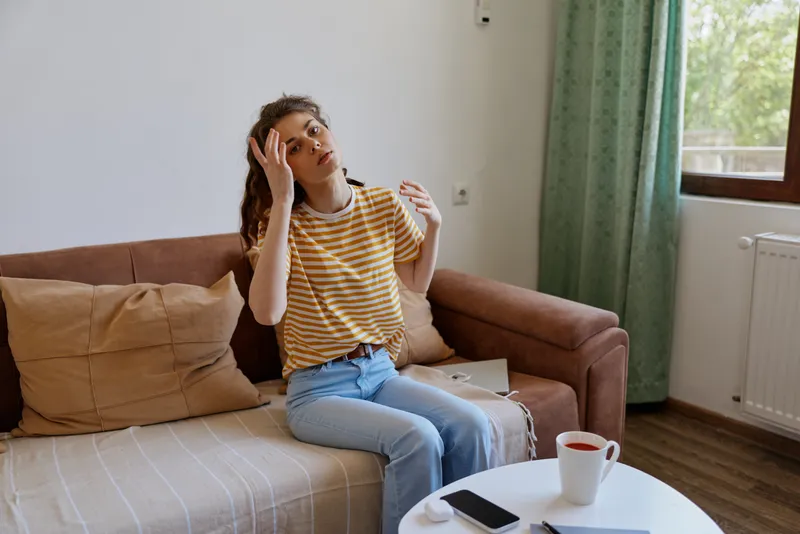 Young woman sits on a beige sofa in a bright living room, touching her hair and looking thoughtful, capturing a casual intimate moment at home today relaxed