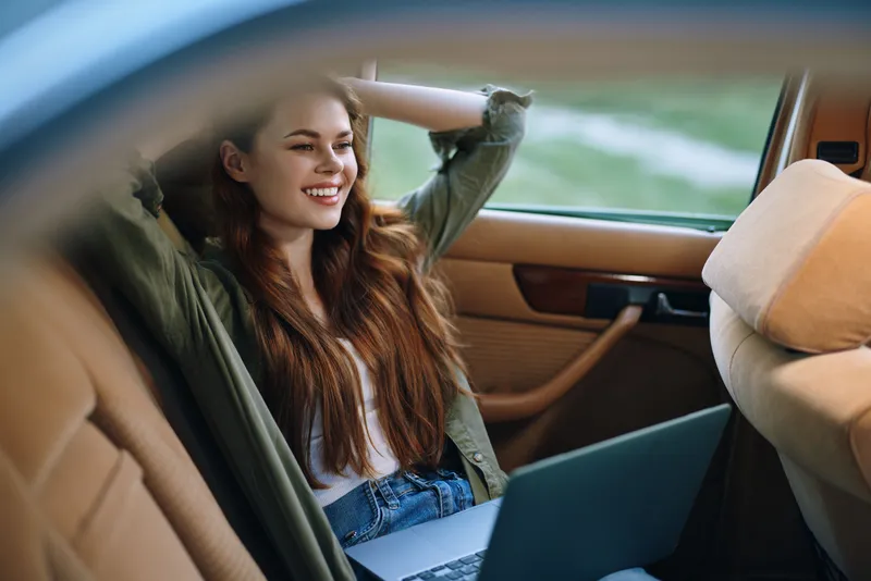 Happy young woman relaxing in a car with a laptop, enjoying the moment on a sunny day, dressed casually in a green jacket, light top, and jeans.