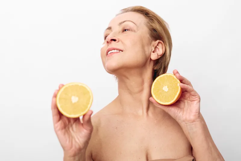 Mature woman with bare shoulders holds two halves of a grapefruit near her face, looking upwards with a serene expression on a white background. Concept of natural beauty and health.