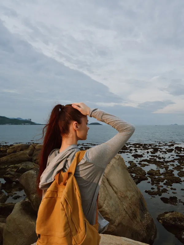 Young woman in a gray hoodie and yellow backpack contemplating by the sea, surrounded by rocks, reflecting calm emotions and nature s beauty