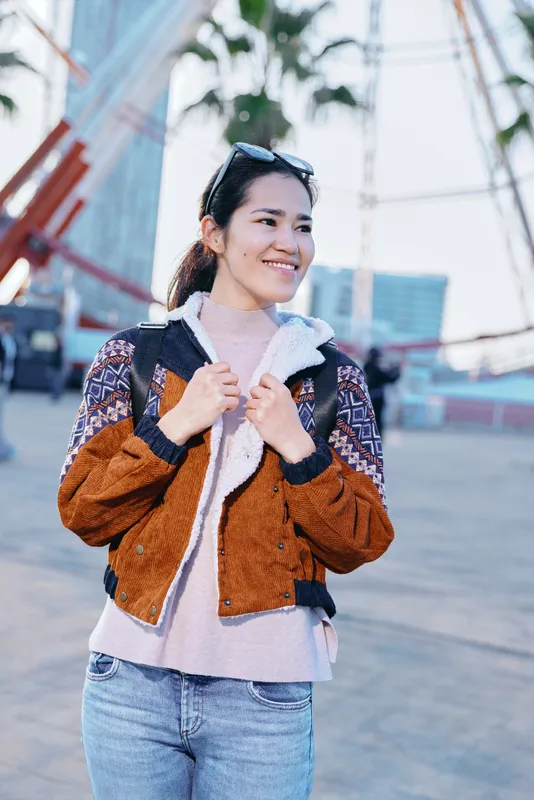 Woman wearing a brown fuzzy jacket and denim jeans, smiling outdoors in an urban plaza, showcasing casual streetwear and confident mood.