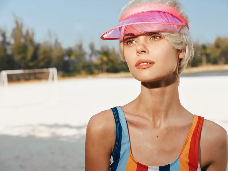 Portrait of a stylish woman in a colorful striped swimsuit and pink visor on a sunny beach volleyball setting, looking forward with confident, serene expression.