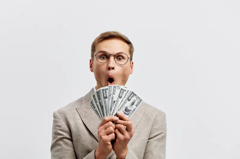 Young surprised man with glasses holding fan of hundred dollar bills, studio shot against plain white background, financial success concept, money, wealth, dollar bills, expression of excitement, pros