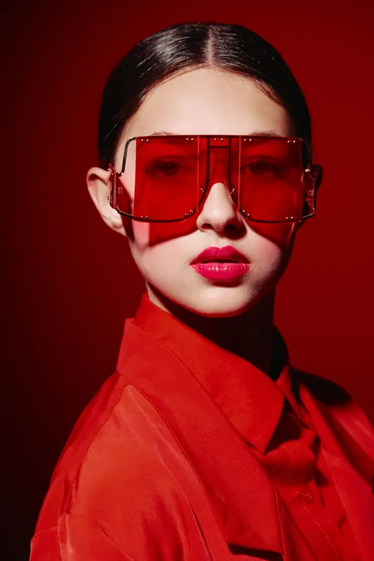 Stylish woman wearing red oversized sunglasses and vibrant red makeup poses against red background. Fashion concept focuses on beauty, creativity, and color coordination.