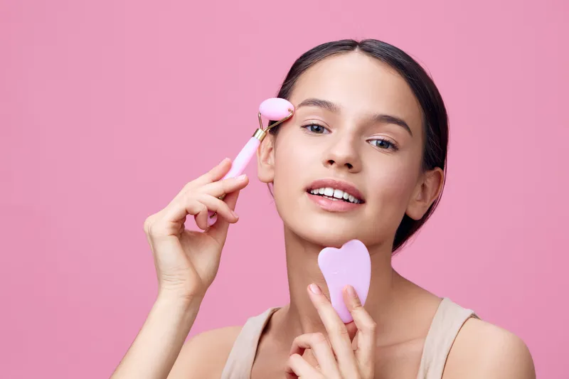 Young woman using facial rollers on pink background, showcasing skincare routine, beauty, and self care with emotional confidence and relaxation