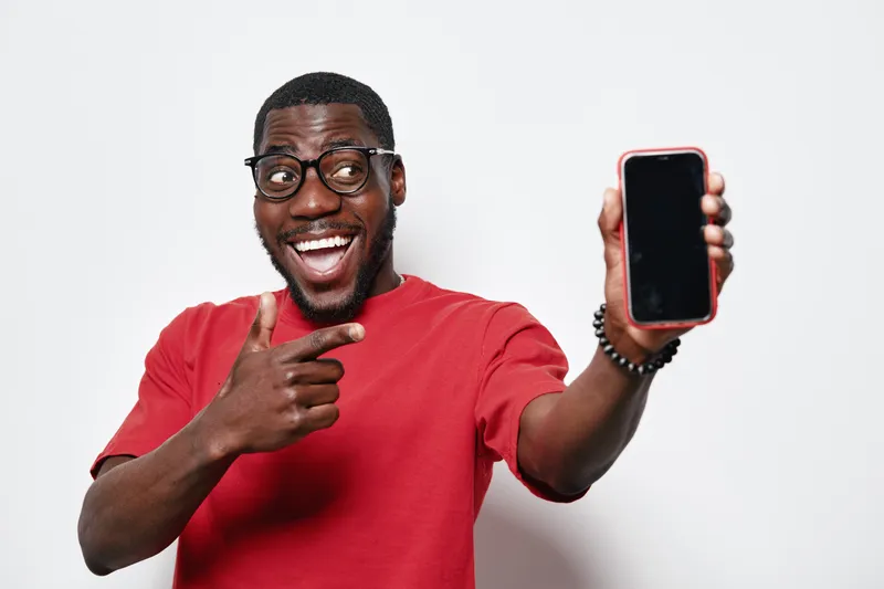 African American man with dark skin wearing glasses and red shirt smiling and pointing at smartphone screen for technology and communication concept on white background.