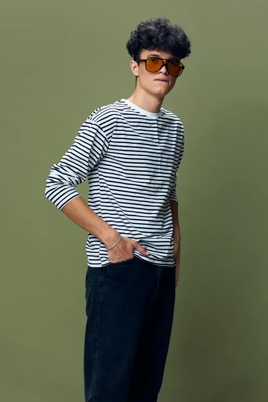 Young man wearing striped shirt and sunglasses stands confidently against olive green background with casual pose and curly hair, displaying youthful style and attitude.