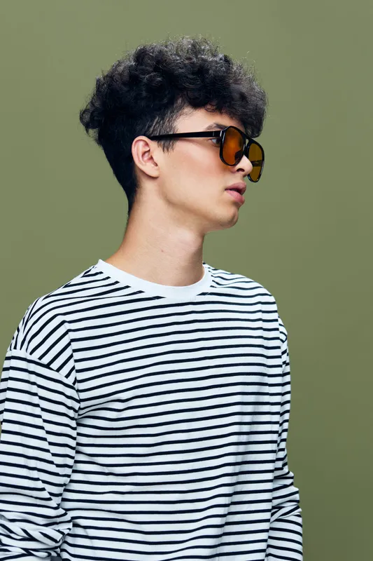 young man wearing sunglasses and striped shirt posing against green background with neutral expression and short curly hair, casual style and modern fashion details
