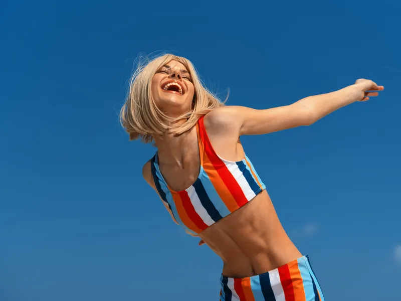 Smiling woman in colorful striped outfit enjoying sunny day outdoors with clear blue sky and joyful energy, celebrating happiness and freedom.
