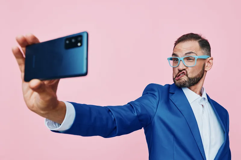A business professional man captures a selfie against a solid pink background, exuding confidence and playful energy while posing with a modern smartphone.