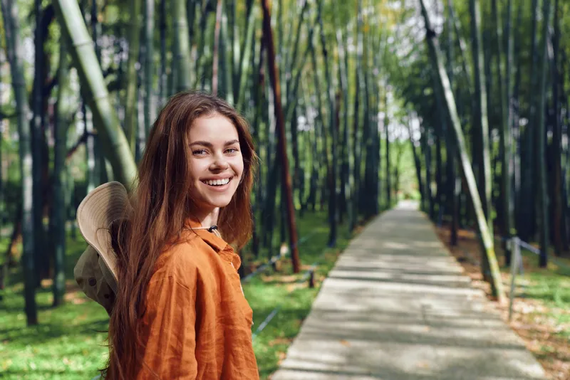 Woman smile bamboo path hat portrait outdoor forest scene with young brunette in casual clothing and straw hat, walking on walkway among green bamboo grove, relaxed travel and nature mood