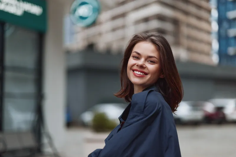 A smiling young woman with long brown hair wears a dark jacket outdoors in an urban setting, looking confidently at the camera during daytime.