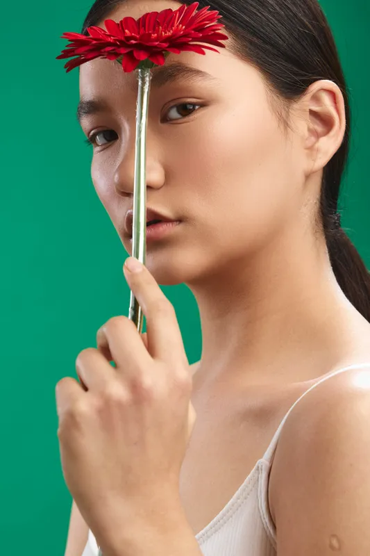 asian woman holds a red flower near her face in a studio portrait, soft lighting and clean background create an elegant, calm and artistic mood with contemporary flair.