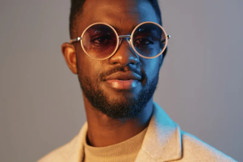 Young Black man wearing stylish round sunglasses, light grey suit jacket, and beige turtleneck, showcasing confidence against a soft blue and orange background