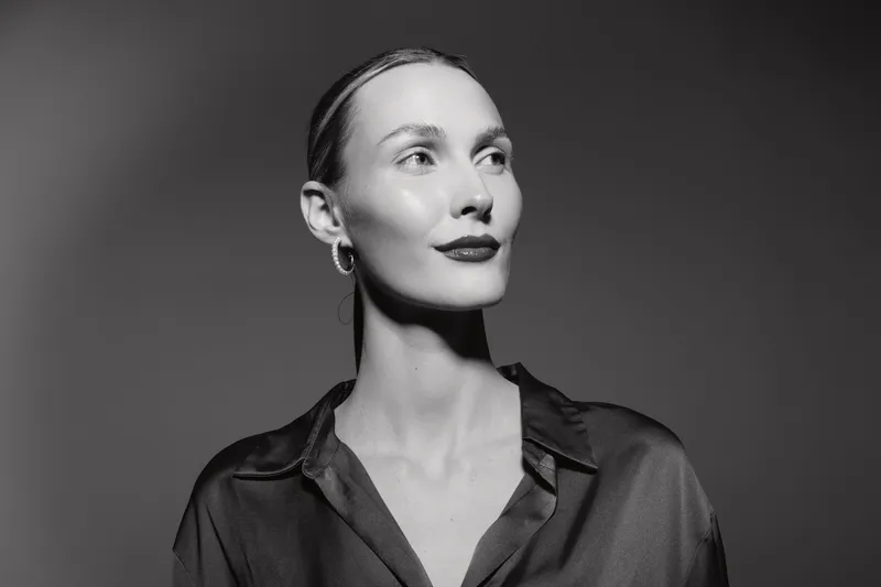 Portrait of a fair skinned white woman with slicked back hair wearing hoop earrings and a dark shirt looking aside in studio lighting.
