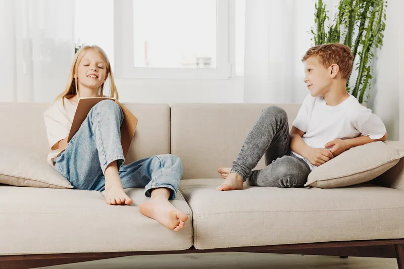 Two children sitting on beige sofa in bright living room, girl reading book and boy watching, casual home lifestyle concept.