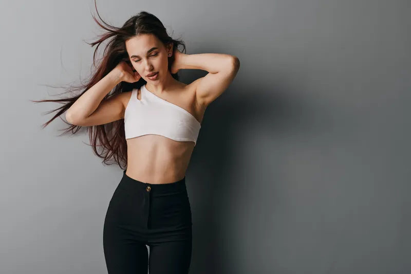 Young woman with long hair wearing white crop top and black high waist pants posing with hands behind head against gray wall in studio. Casual style and natural look for fashion portrait.