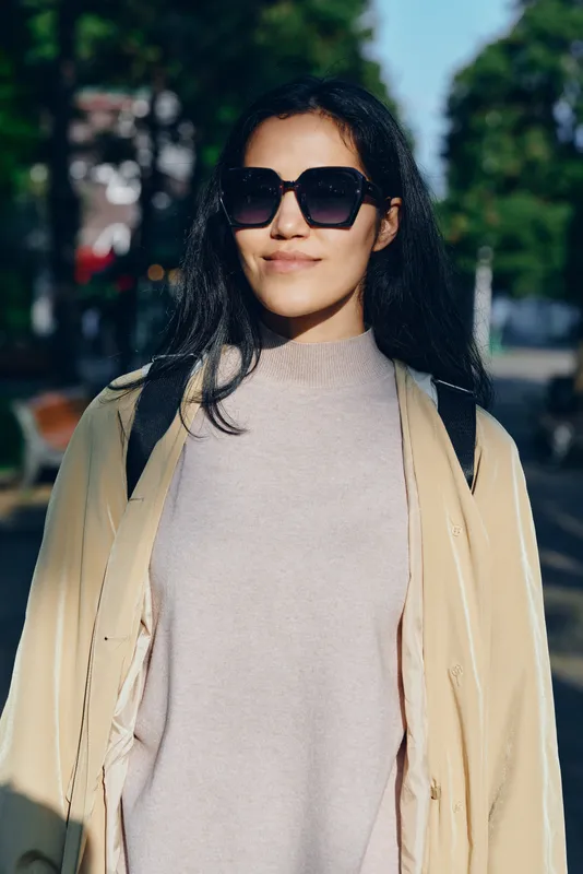 Sunglasses worn woman walking city street outdoors in daylight, casual fashion, smiling softly in light coat and sweater, urban vibe with trees in the background