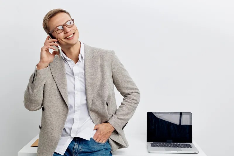 Smiling woman talking on mobile phone with laptop on table, casual office environment, cheerful expression, glasses, light gray blazer, white shirt, modern workspace, professional mood, bright lightin