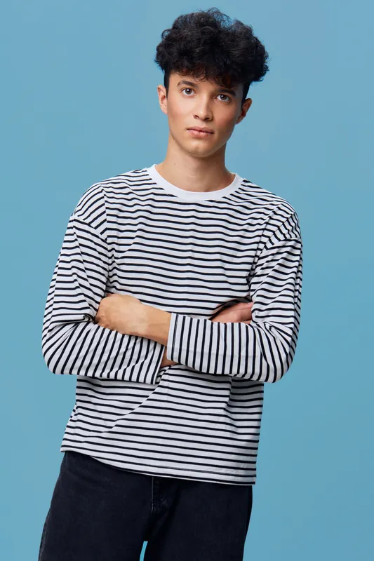 Young man wearing a striped long-sleeve shirt stands with arms crossed against a plain blue background, showcasing casual style and relaxed confidence.