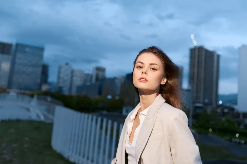 confident woman in beige jacket standing outdoors in cityscape with cloudy sky and modern buildings at dusk KEYWORDS: confident woman, woman, cityscape, businesswoman, modern buildings, beige jacket, 