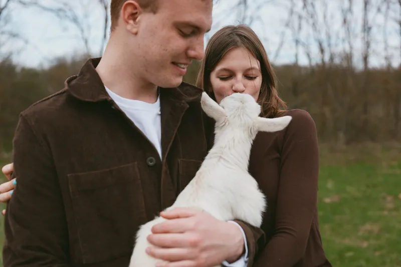 A couple outdoors with a gentle goat kid, sharing affectionate moment in a rustic countryside setting, showcasing lifestyle, tenderness, bonding, and calm, as the animal nuzzles close.