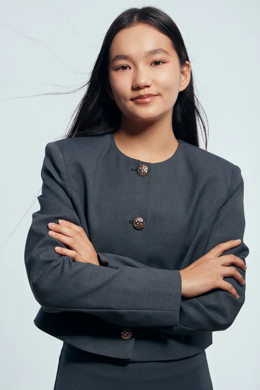 Young businesswoman standing with arms crossed wearing dark formal suit with decorative buttons looking confident and professional against a plain background.