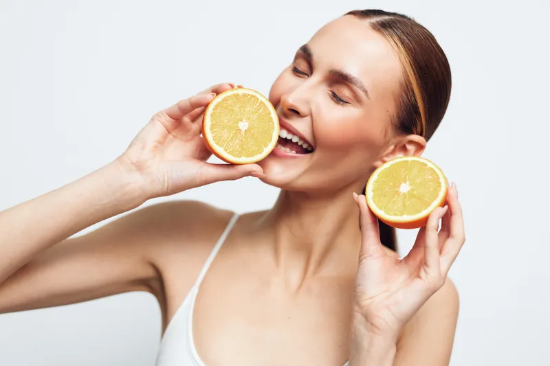 Woman holds orange slices and eats citrus with a smile, suggesting vitamin healthy nutrition, wellness routine, skincare fresh juice in soft studio lighting