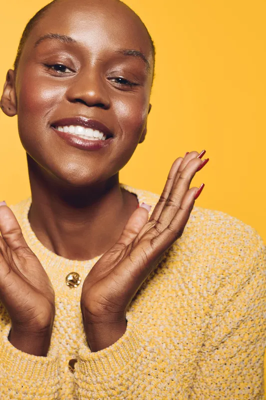 Smiling black woman with dark skin wearing yellow sweater poses happily with hands near face. Bright yellow background highlights joyful mood and natural beauty of African ethnicity.