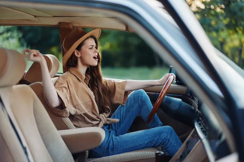 Young woman enjoying a road trip, wearing a beige hat and casual attire, expressing joy while driving in a vintage car amidst a green landscape.