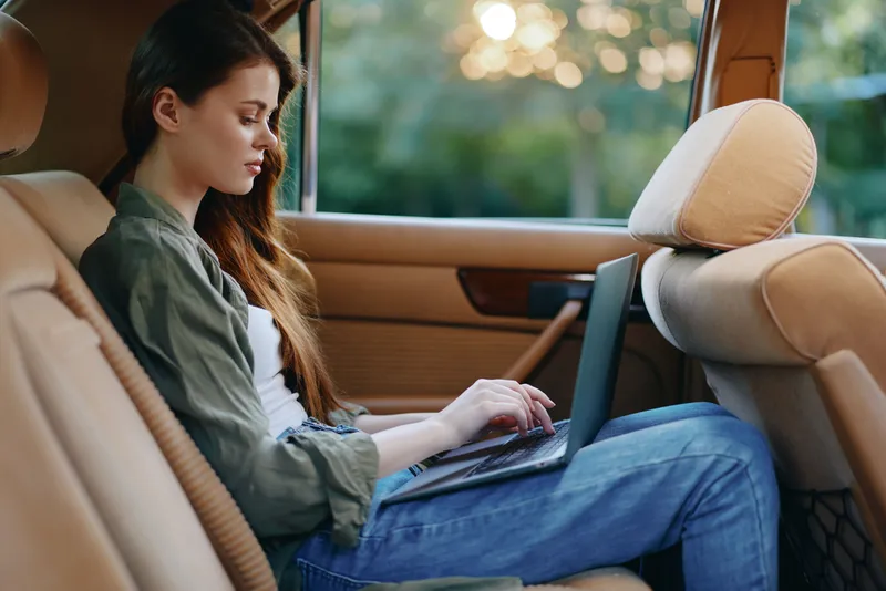 Young woman working on a laptop in the backseat of a car, wearing casual clothes with a calm expression, surrounded by warm natural light.