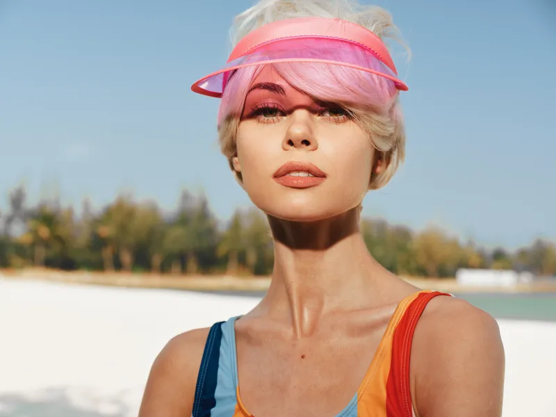 Closeup portrait of a stylish woman wearing a pink visor on a sunny beach, radiant skin, bold colors, and a confident expression under bright coastal light.