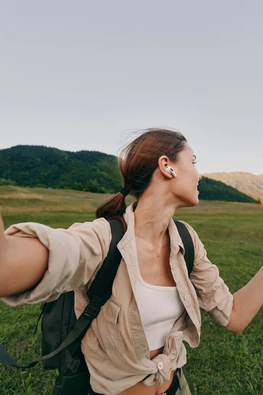 Cheerful woman outdoors, backpacker in beige shirt and white top, reaching toward camera, sunlit meadow scene, rolling hills, natural colors, relaxed lifestyle moment