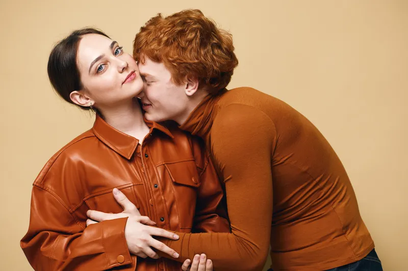 Couple in brown clothing hugging and showing affection on beige background with woman looking calm and man whispering or kissing near her face in indoor studio. Intimate moment and close emotional