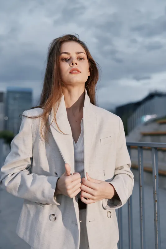 confident woman in beige jacket standing outdoors with urban background at cloudy weather, fashion portrait of young European woman