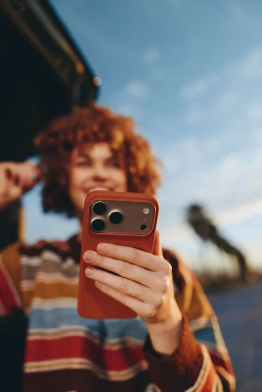 Woman lifestyle outdoors, curly redhead smiling, holding smartphone for selfie, casual striped sweater, warm daylight, soft breeze, shallow depth of field, friends walking behind