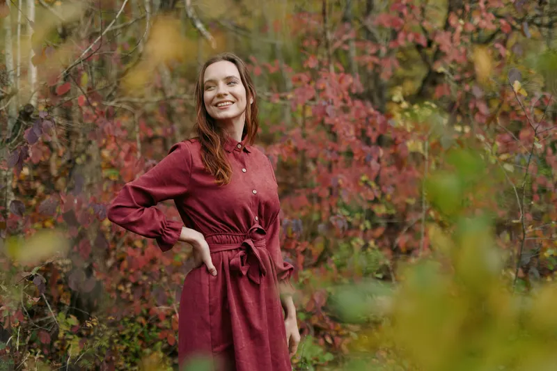 A smiling young woman in a burgundy dress stands among autumn foliage, captured in a candid natural moment that highlights authenticity and a genuine emotional presence in the frame.