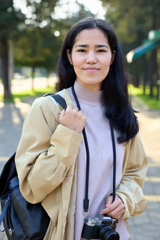 Camera in hand, a woman stands in a sunlit park wearing a beige jacket and backpack, ready for outdoor photography and exploration, capturing casual travel moments, nature, and everyday life.