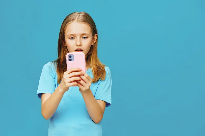 Young girl surprised while using smartphone, wearing blue t shirt, against vibrant blue background, expressing curiosity and amazement