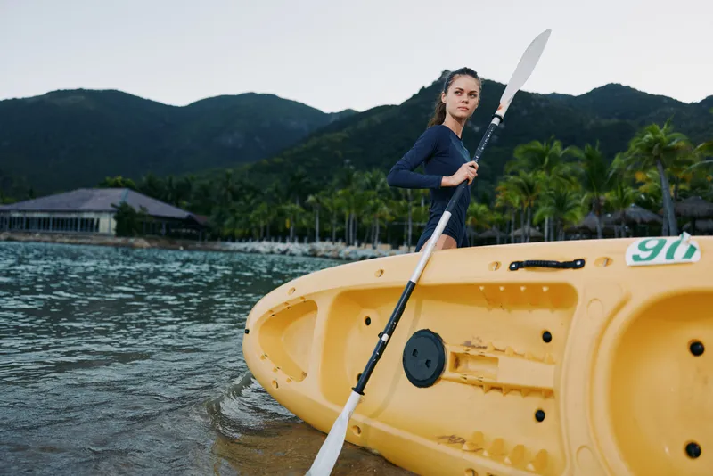 A fit young woman stands with a paddle beside a bright yellow kayak on a tropical shoreline, ready for a water adventure under a calm, clear sky.