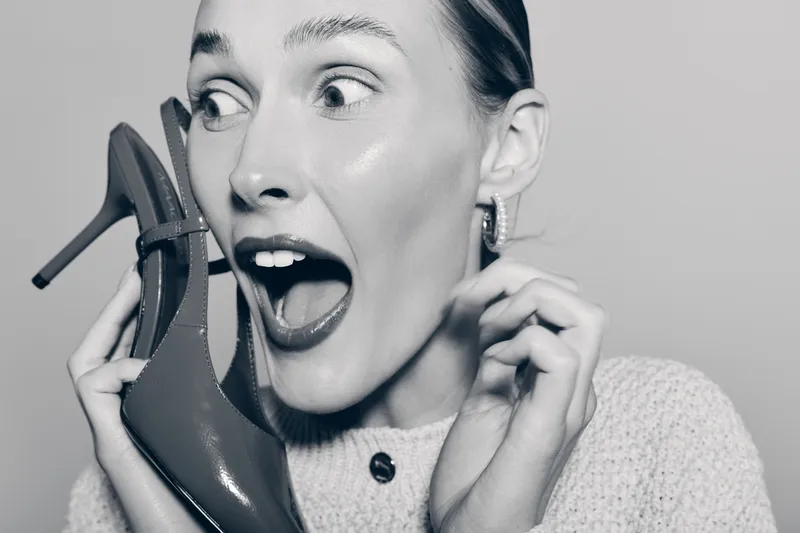 Young white woman with light skin holds high heel shoe near face with excited expression in black and white photo concept of fashion and emotion.