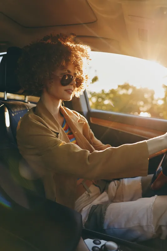 A woman driver with curly hair and sunglasses sits behind the wheel of a vintage car, capturing a relaxed lifestyle moment with warm retro film colors.