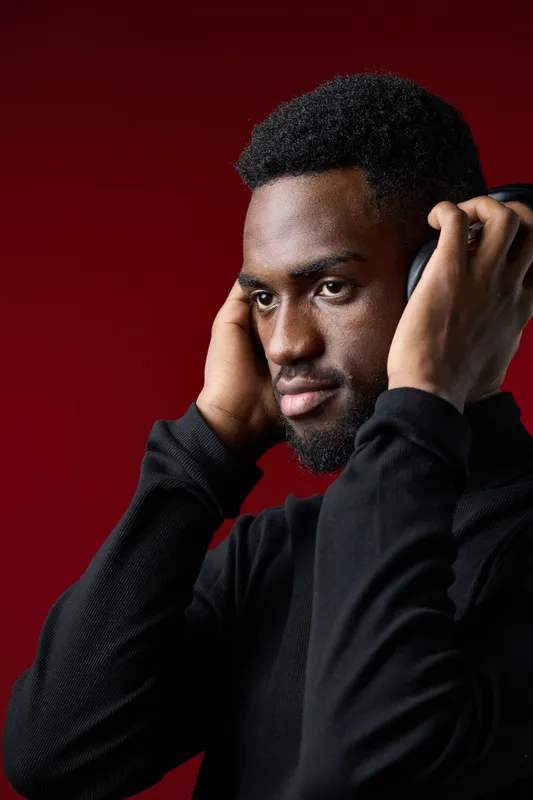 Portrait of a stylish young Black man wearing headphones, expressing focus and relaxation against a vibrant red background, ideal for music and lifestyle concepts